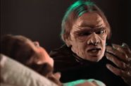 Man with wrinkles on his face speaks to a woman who is lying down.