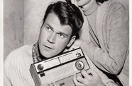 A man sits holding a radio, while a woman leans on him with a shocked expression.
