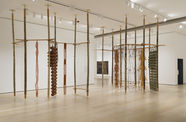 Gallery space with several long, wooden poles that reach from the floors to the ceilings adorned with woven strings and cylinders. 