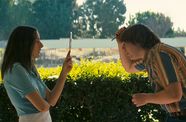 Girl and boy stand facing each other in front of a sunny hedge.