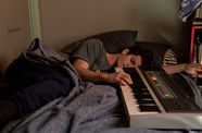 A man lies down on a bed with his fingers on a piano keyboard.