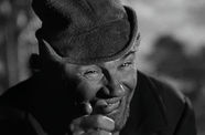 A man in a hat points at the camera. 