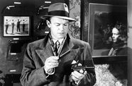 A man in a suit and fedora holds a pistol.