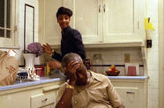 Man sits at a kitchen table, slumped over, while a woman is behind him. 