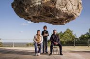 Film still from "Official Competition" (2022), showing a gigantic rock suspended over three people who seem not to notice