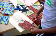 A table of scrap paper with different patterns and a kid standing to the right cutting one heart-shaped image for a collage.