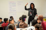 A woman with brown skin wearing all black holds her right arm up in front of a group of young children seated at tables covered in kraft paper.