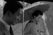 A man and woman hold umbrellas while looking at the river