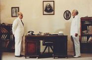 two men in white suits stare at each other across the length of a wooden desk