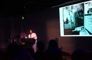 Speaker at a podium looking toward an image projected on a screen