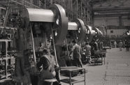 Men work on large metal machines. Black and white still.