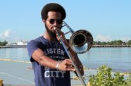 A Black man with an afro plays a trombone in front of a river