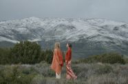 Two brightly dressed women stand facing each other in a field with a snow-capped mountain behind them