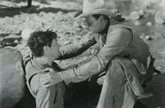 man with cowboy hat holds the shoulders of another man leaning against rock