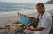 Rogê sitting with a guitar on a beach