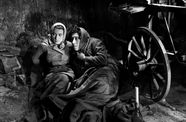 A man and woman with scarves on their heads huddle next to a carriage wheel