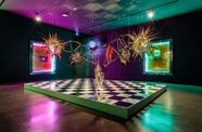 View of a gallery lit in green and pink, with a large, vibrant sculpture hanging from the ceiling