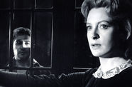 A woman stands in the foreground and a man in the background, separated by prison bars