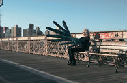 Artist Sylvia Palacios Whitman sitting on a bench on the Brooklyn Bridge, wearing an oversized hand