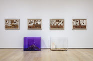 Framed newspaper sheets with words cut out hang on a wall, with two lucite boxes full of paper scraps sit in front