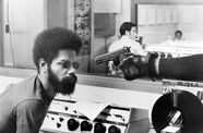 A gloved hand points a handgun at a Black man with a beard and afro