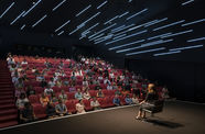 An audience sitting in a darkened theater, watching a woman on stage