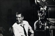 A smiling Desi Arnaz, sitting next to a film camera, looks out of frame