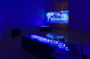 Installation with a video screen in the background showing a street scene, and a neon sign reading "til' somebody" laying on a plinth in the foreground