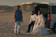 A couple dressed for their wedding sits in the open hatch of a van in the dessert