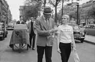 A man and a suit and a woman in a New York Herald Tribune shirt walk down a city street, followed by a cameraman