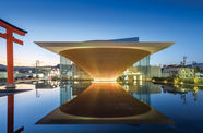 A glowing building in the shape of an inverted pyramid, with a reflecting pool in front of it