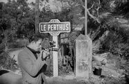 A man loks through a small, handheld film camera as he stands in front of a sign reading "LE PERTHUS"
