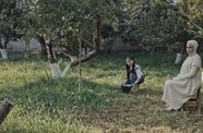 A girl kneels next to a woman in all white amidst a garden