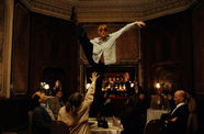 Dancer with red bangs stands on a restaurant dining table lifting their right leg high in the air, as the people sitting at the table reach up at them