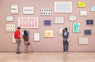 Three visitors, viewed from behind, stand in front of a pink gallery wall covered in a cluster of framed abstract artworks
