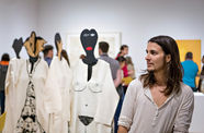 A person with long, straight brown hair and a short beard glances to the right. Just behind him are visible four mannequins wearing caftans with designs painted onto them