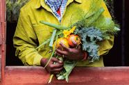 Person in a mustard coat and patterned scarf holds a colorful harvest of carrots, squash, onions, and leafy greens through a red window.
