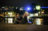 Two women sit on the waterfront, with city lights twinkling behind them