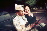 A woman in a black dress and a man in a solider's uniform sit in the front seat of a jeep, the man holding the steering wheel while the woman rests her elbow on his shoulder and smiles