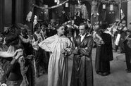 Two men in silken robes stand in the middle of a hall filled with dancing couples