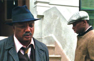 Two men, one facing the camera wearing a fedora and suit, the other facing down