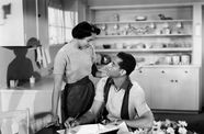 A woman stands over a man sitting at a table, pencil in hand, in a kitchen