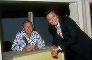 A man in a pin-striped suit smiles as he leans against a desk, where another smiling man sits in front of a microphone