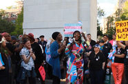 A dark skinned person with long black hair and a colorful dress speaks into a loudspeaker at an outdoor protest