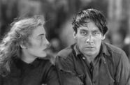 On the right, a man with dark hair swept across his forehead looks at the camera, while a woman on the left gazes at him