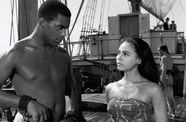 A man and woman looking at each other on a ship. The man is enslaved in shackles.