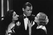 A dapper man in a tuxedo looks at a blond woman clinging to his left arm, while a dark haired woman clings to his right