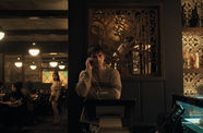 A young man in a striped sweater holds a cell phone next to his head, while out-of-focus in the background, a long-haired figure in a beige sweater and dark skirt looks his way