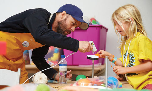 A girl in a yellow shirt is holding up an art project and is assisted by a man in a baseball cap who leans over with a glue gun pointed at the girl's project.