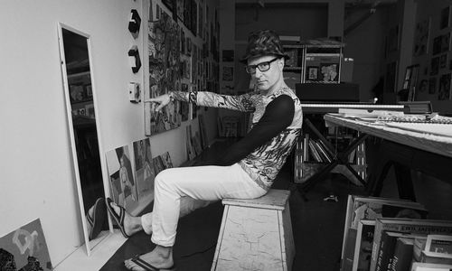 Black and white photo of artist Wayne Koestenbaum seated and pointing at himself in a mirror while looking at the camera